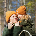 Nainen kantaa lasta reppuselässä auringon paistaessa metsäisellä taustalla. Molemmilla on päässä oranssit pipot.