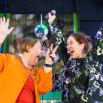 Kaksi iloisesti hymyilevää naista nostavat kädet ylös ja pitelevät toisessa kädessä mikrofonia.