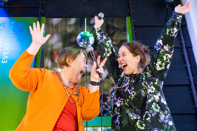 Kaksi iloisesti hymyilevää naista nostavat kädet ylös ja pitelevät toisessa kädessä mikrofonia.