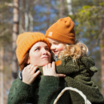 Nainen kantaa lasta reppuselässä auringon paistaessa metsäisellä taustalla. Molemmilla on päässä oranssit pipot.