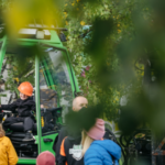 Lapsi istuu oranssi kypärä päässä metsäkoneessa leveästi hymyillen. Neljä muuta henkilöä katselee vihertävässä ympäristössä ympärilleen.