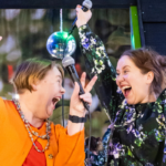 Two women laughing happily, holding hands up in front of a disco ball. In the other hand, both have a microphone.